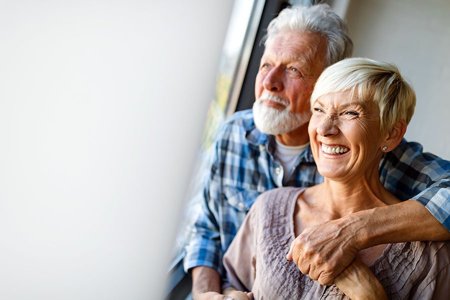Medicare - Happy Senior Couple Hugging and Laughing While Looking Out the Window of Their Home